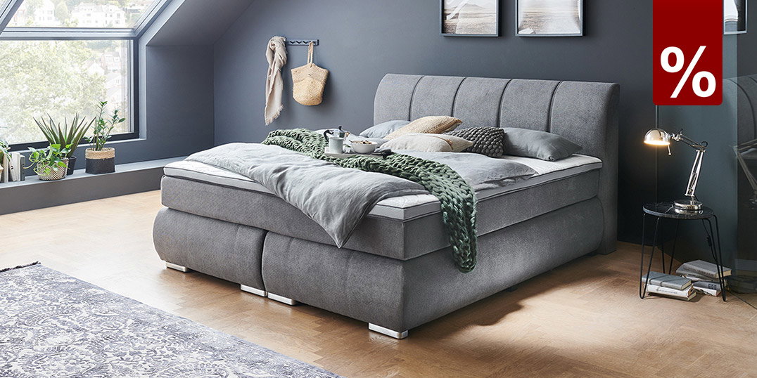 Modernes Boxspringbett in Anthrazit mit Topper, Kissen und Bettwäsche in einem offenen Schlafzimmer vor dunkler Wand