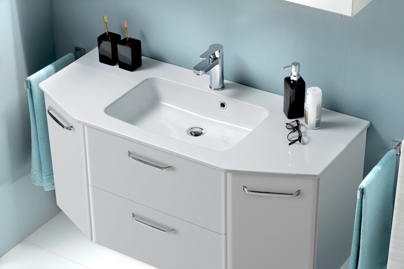 Moderne witte wastafel met geïntegreerde onderkast en chromen kraan, voorzien van een zeepdispenser en tandenborstelbekers, afgebeeld vanuit een schuin vooraanzicht van bovenaf.