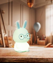 Niedliches Akku-Nachtlicht in Form eines Hasen mit Brille, frontal auf einem Tisch in einem gemütlichen Raum platziert.