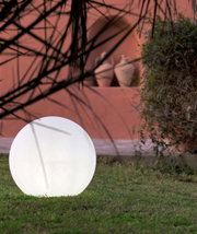 Een stralende, bolvormige buitenlamp in het wit op een grasveld voor een terracottakleurige muur, gefotografeerd vanuit een laag standpunt bij de grond