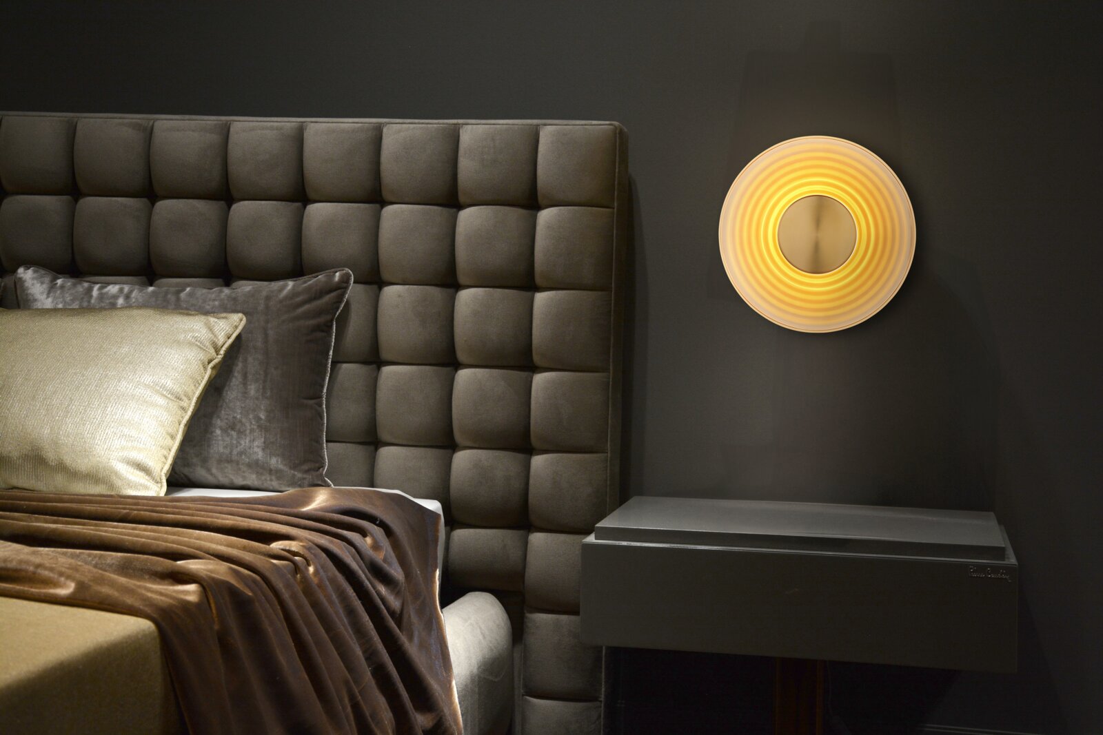 Moderne runde Wandleuchte mit warmem, konzentrischem Lichtschein über einem Nachttisch neben einem gepolsterten Bettkopfteil, frontal aus Augenhöhe fotografiert