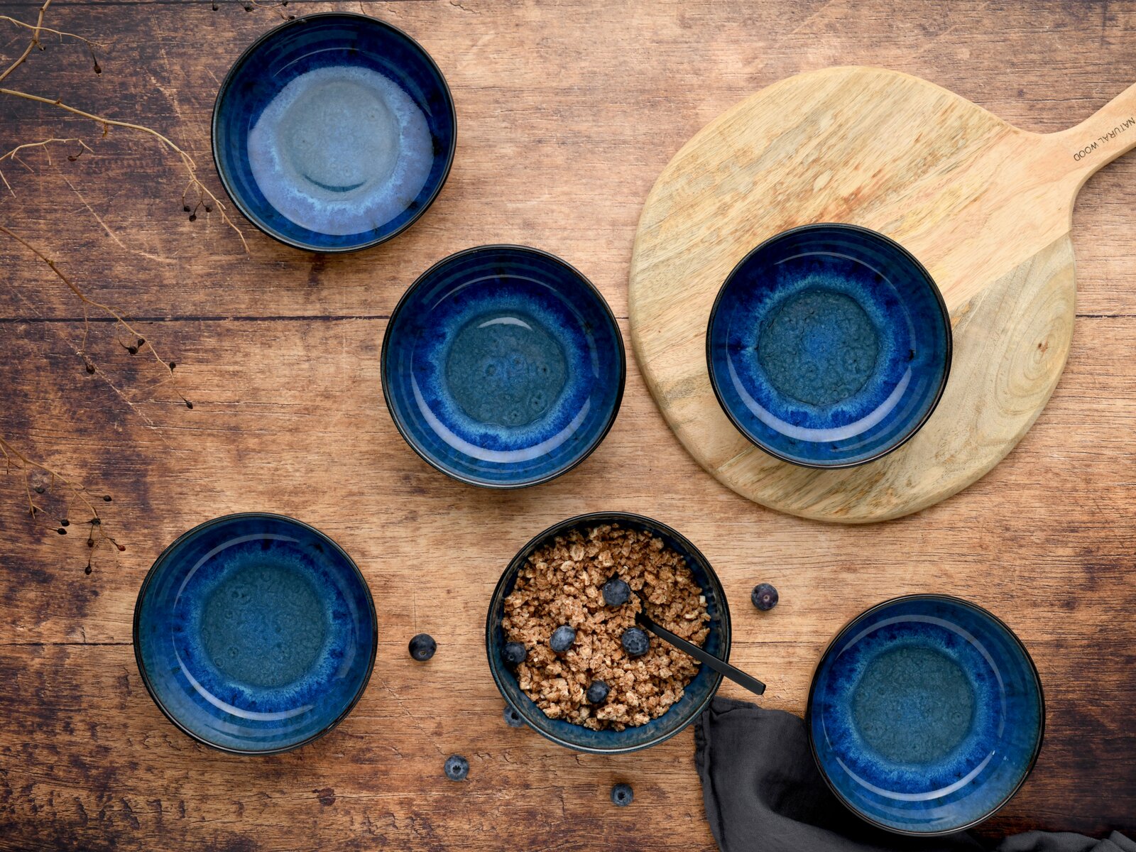 Dunkelblaue Keramikschalen mit reaktiver Glasur auf rustikalem Holztisch, eine Schale mit Müsli und Blaubeeren, daneben Holzbrett; Draufsicht von oben