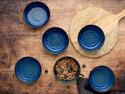 Dunkelblaue Keramikschalen mit reaktiver Glasur auf rustikalem Holztisch, eine Schale mit Müsli und Blaubeeren, daneben Holzbrett; Draufsicht von oben