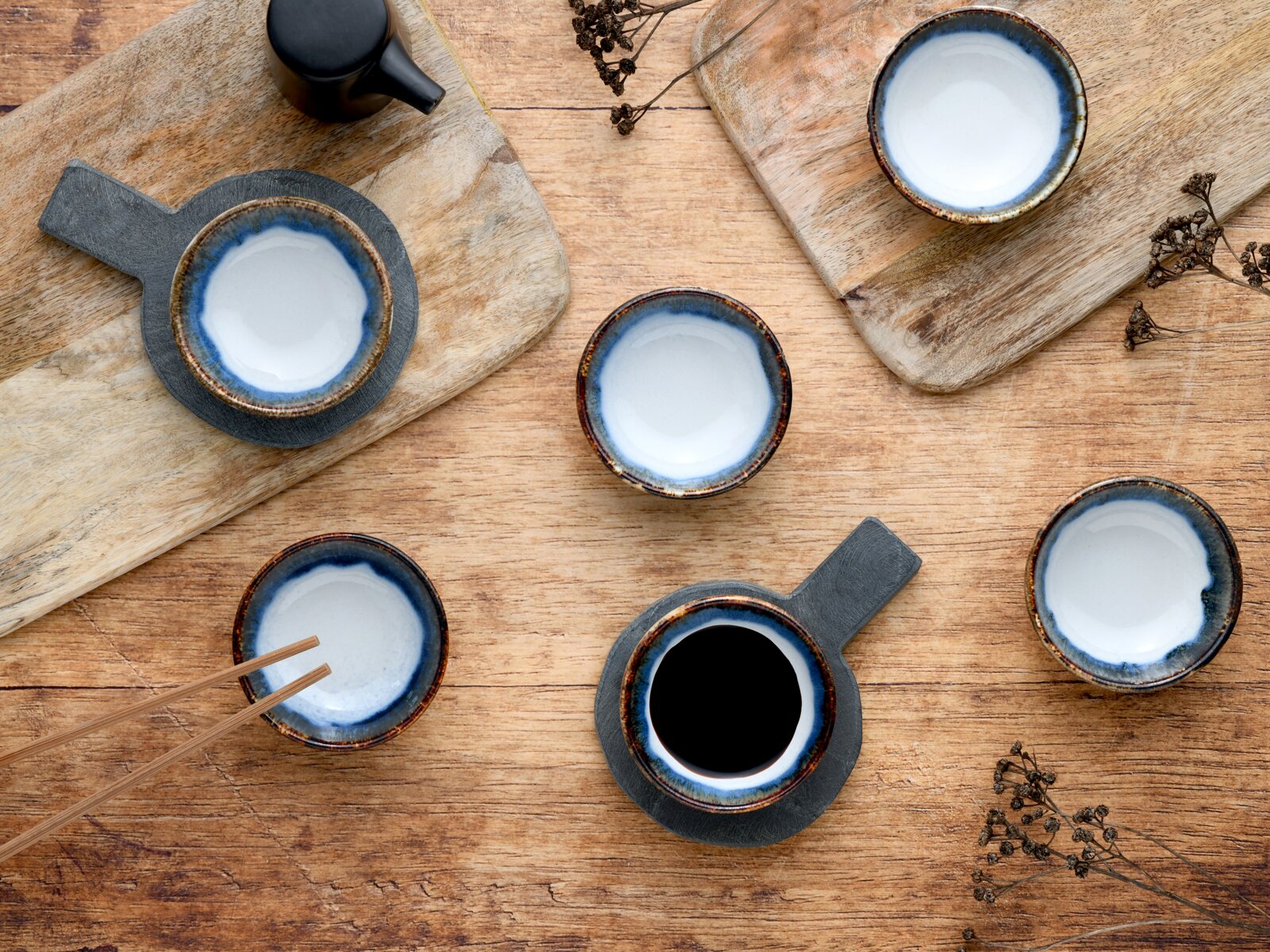 Draufsicht auf mehrere handgefertigte Keramikschalen und Tassen mit blauem Rand auf rustikalem Holztisch, teils auf Holz- und Schieferbrettern, eine Tasse mit schwarzem Kaffee.