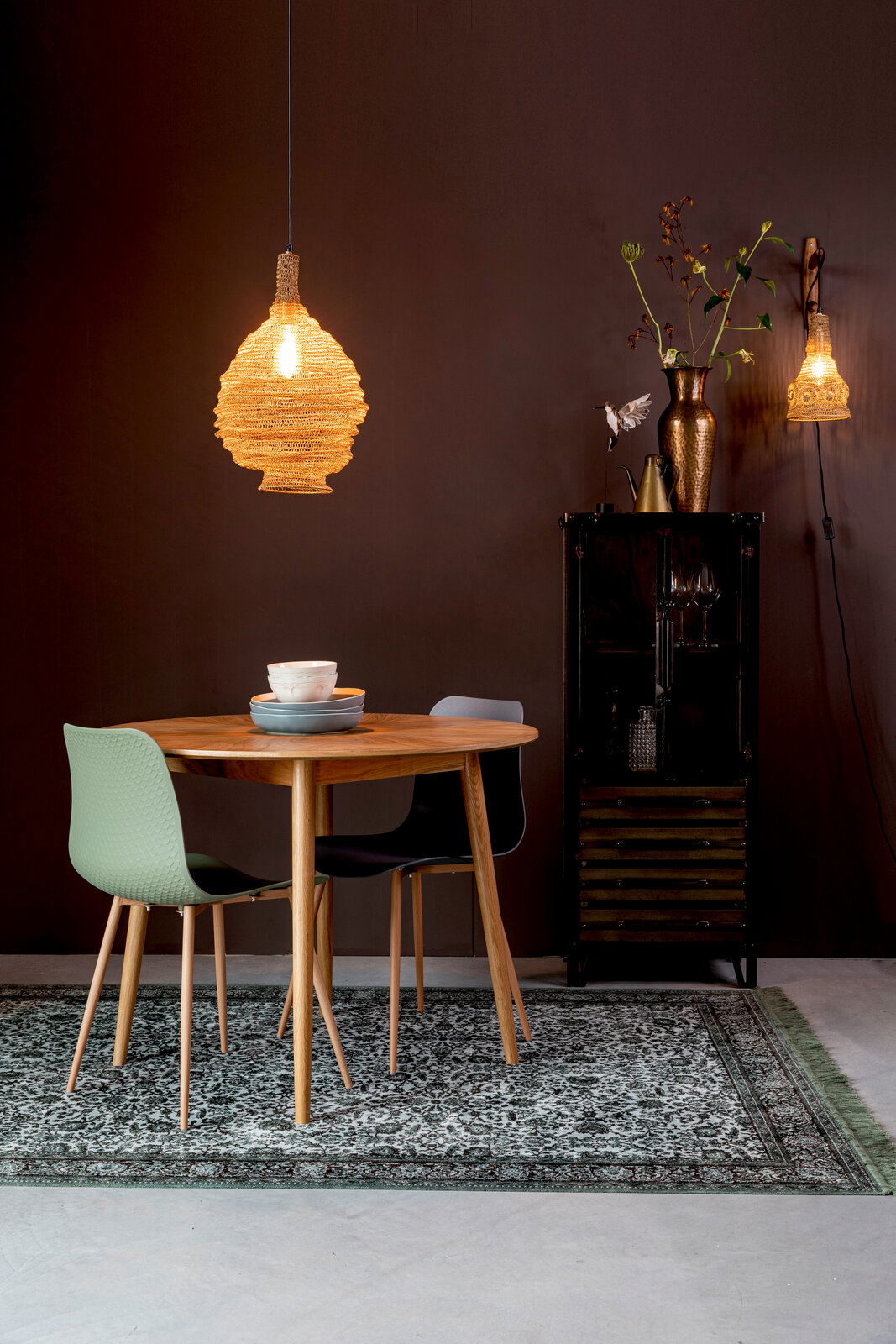 Runder Esstisch aus Holz mit zwei modernen Stühlen, einer in Grün und einer in Schwarz, auf einem gemusterten Teppich. Im Hintergrund eine stilvolle Hängelampe und eine dekorative Vitrine. Perspektive: Frontalansicht.