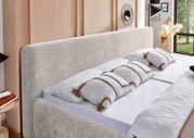 Modernes Doppelbett mit hohem, weich gepolstertem Kopfteil in hellem Beige, aus schräger Vorderansicht in einem stilvollen Schlafzimmer gezeigt