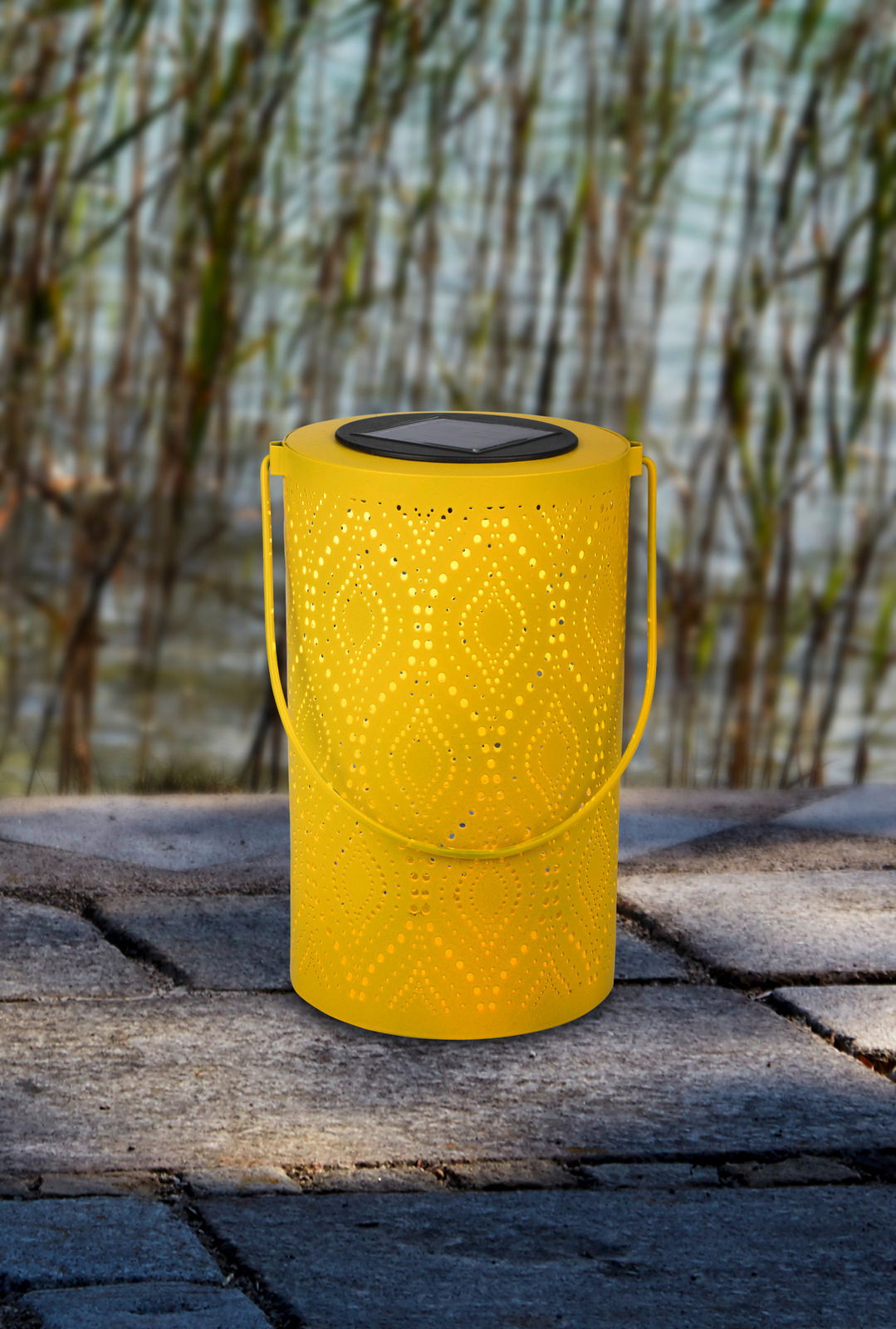 Gelbe Solarleuchte mit dekorativem Muster, seitliche Perspektive, auf einem Steinboden vor Schilf im Hintergrund.