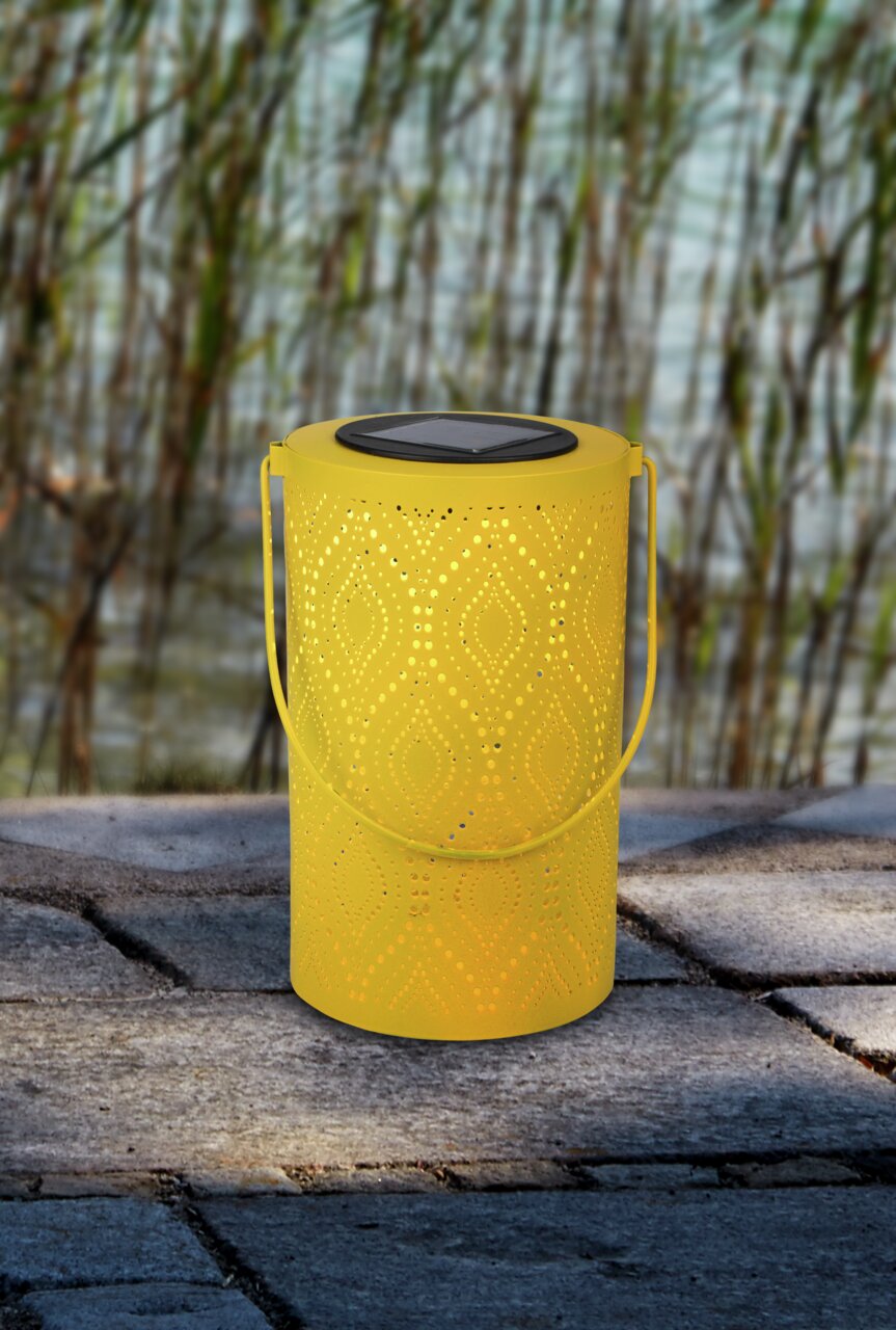 Gelbe Solarleuchte mit dekorativem Muster, seitliche Perspektive, auf einem Steinboden vor Schilf im Hintergrund.