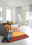 Bunte Decke mit geometrischem Muster in warmen Farben, drapiert über einem grauen Sofa in einem modernen, hellen Wohnzimmer. Perspektive von vorne.