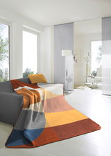 Bunte Decke mit geometrischem Muster in warmen Farben, drapiert über einem grauen Sofa in einem modernen, hellen Wohnzimmer. Perspektive von vorne.