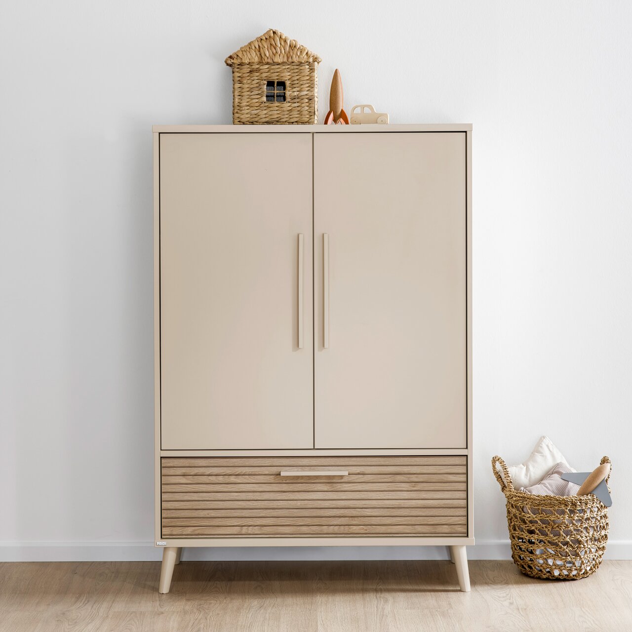 Moderne kledingkast met twee deuren en een lade, gefotografeerd vanaf de voorkant. Bovenop staan een decoratief mandhuisje en een klein speelgoedautootje. Ernaast staat een gevlochten mand met kussens.