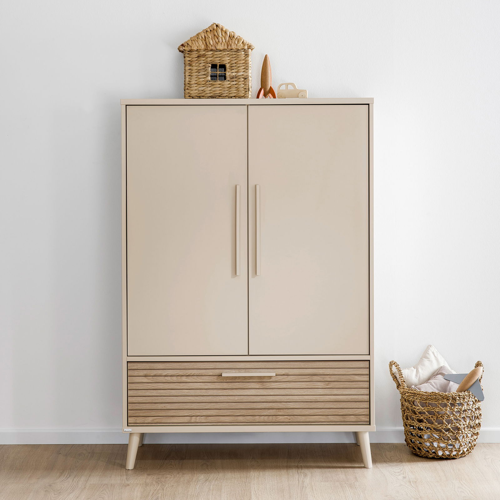 Moderner Kleiderschrank mit zwei Türen und einer Schublade, frontal fotografiert. Obenauf steht ein dekoratives Korbhaus und ein kleines Spielzeugauto. Daneben befindet sich ein geflochtener Korb mit Kissen.