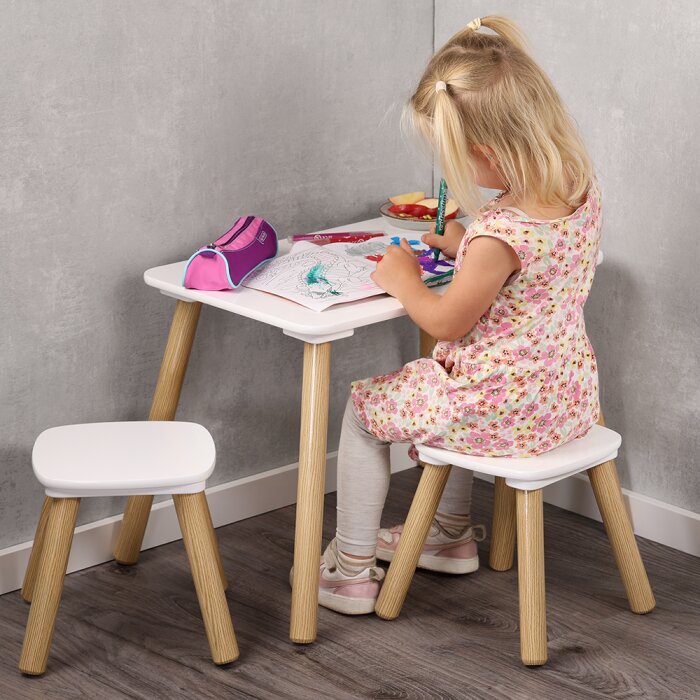 Schräg seitliche Rückansicht: Kleines Mädchen sitzt auf einem weißen Kinderhocker mit Holzbeinen an einem passenden weißen Kindertisch und malt, daneben steht ein weiterer Hocker.