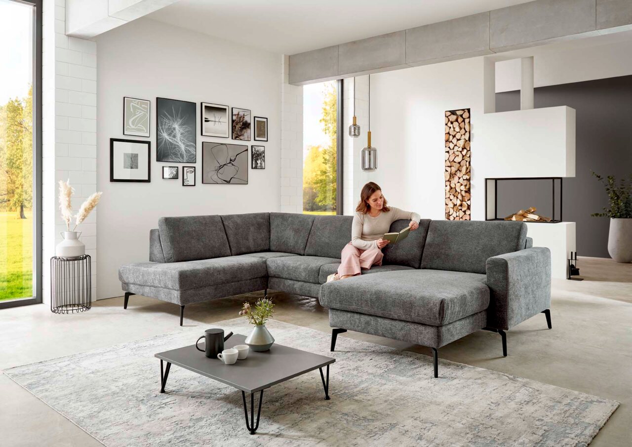 Graue Polsterrundecke in modernem Wohnzimmer, seitliche Perspektive, mit einer Frau, die auf dem Sofa sitzt und ein Buch liest.