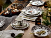 Festlich gedeckter Holztisch mit weiß-braun gemustertem Porzellan-Geschirr (Teller, Schalen und Tasse) mit winterlichem Motiv, goldfarbenem Besteck, grünen und amberfarbenen Gläsern sowie Teelichtern – perspektivisch aus leicht erhöhter, schräger Draufsic