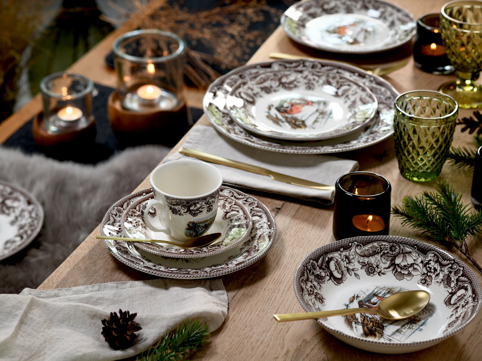 Festlich gedeckter Holztisch mit weiß-braun gemustertem Porzellan-Geschirr (Teller, Schalen und Tasse) mit winterlichem Motiv, goldfarbenem Besteck, grünen und amberfarbenen Gläsern sowie Teelichtern – perspektivisch aus leicht erhöhter, schräger Draufsic