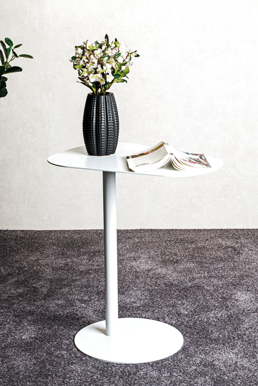 Moderner weißer Beistelltisch mit runder Platte, darauf eine schwarze Vase mit Blumen und ein geöffnetes Magazin, seitliche Perspektive.