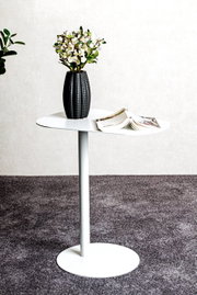 Moderner weißer Beistelltisch mit runder Platte, darauf eine schwarze Vase mit Blumen und ein geöffnetes Magazin, seitliche Perspektive.
