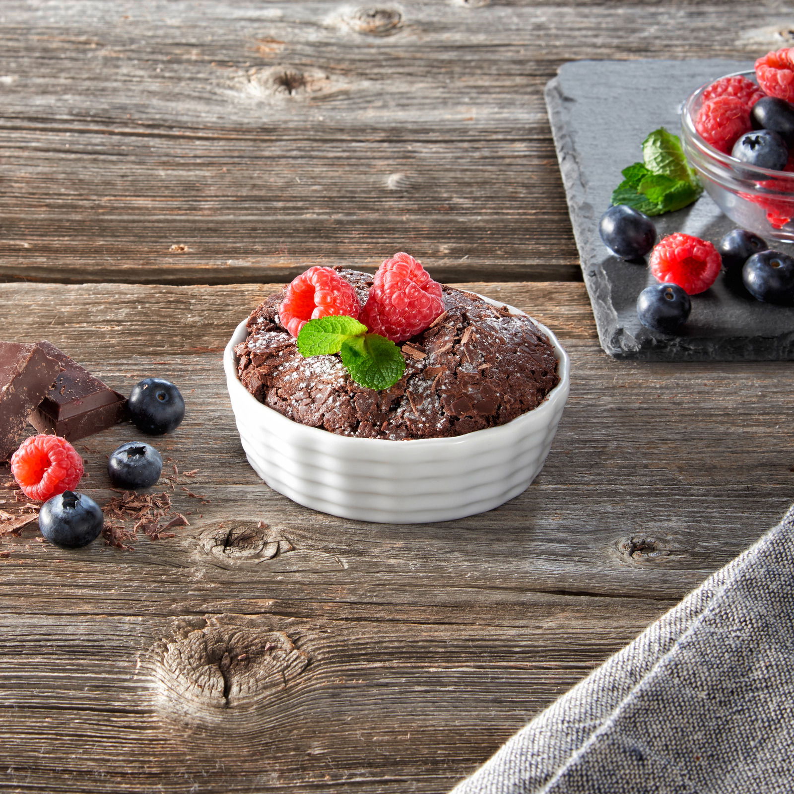 Aufsicht auf eine weiße, geriffelte Auflaufform mit einem Schokoladenkuchen, garniert mit Himbeeren und Minzblättern, auf einem rustikalen Holztisch.