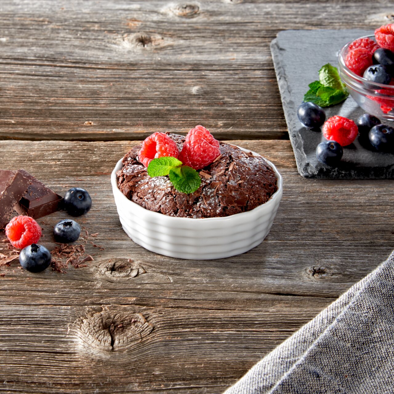 Aufsicht auf eine weiße, geriffelte Auflaufform mit einem Schokoladenkuchen, garniert mit Himbeeren und Minzblättern, auf einem rustikalen Holztisch.