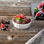Een wit, geribbeld ovenschaaltje met een chocoladetaart, gegarneerd met frambozen en muntblaadjes, op een rustieke houten tafel.
