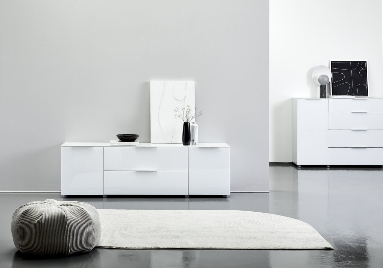 Modernes weißes TV-Lowboard mit Hochglanzoberfläche, frontal fotografiert, dekoriert mit einer Vase und einem Kunstwerk.