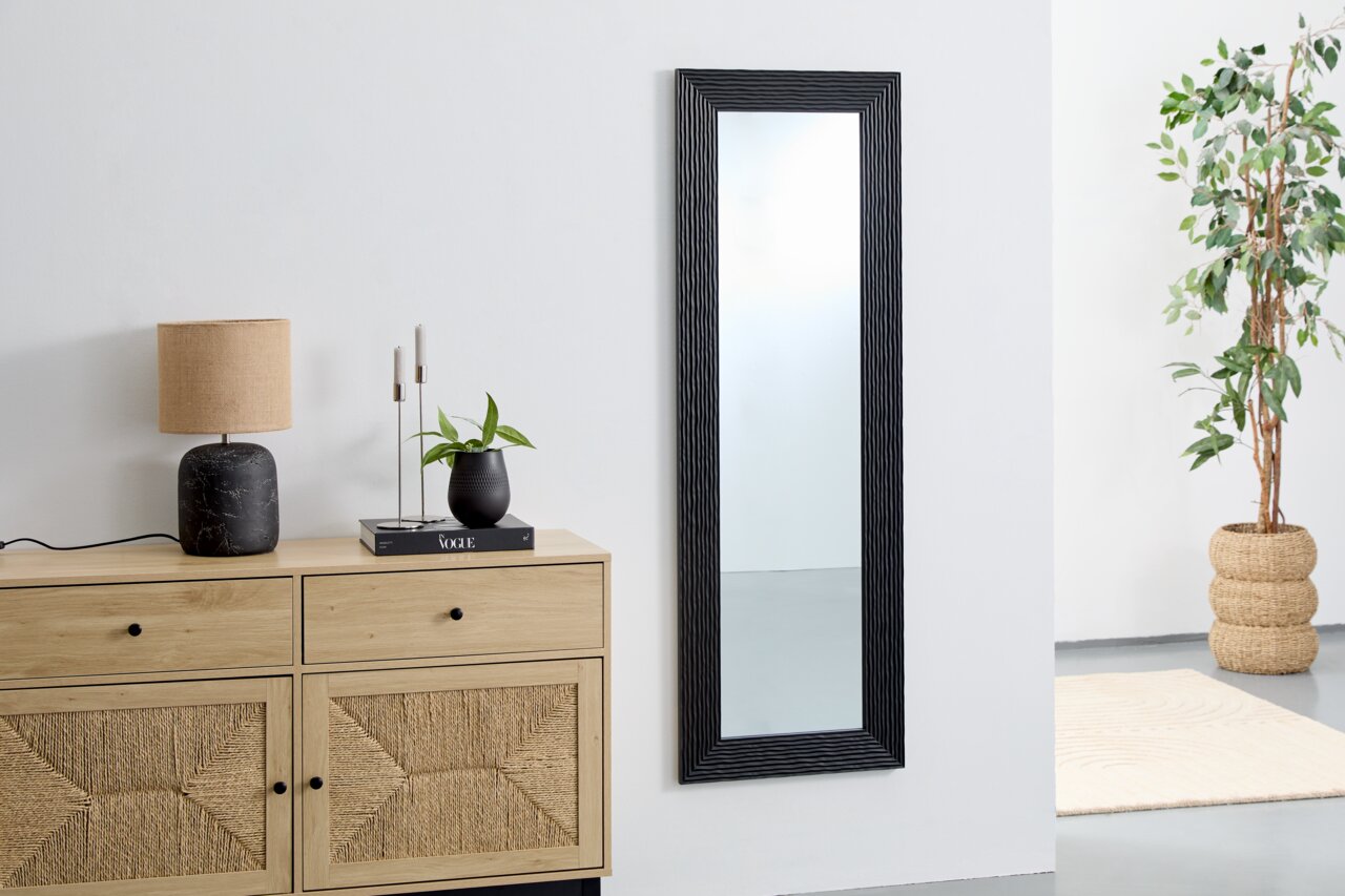 Schwarzer Wandspiegel mit geriffeltem Rahmen, ca. 50x150 cm, in einem modernen Raum mit Kommode, Lampe und Pflanze, seitliche Perspektive.