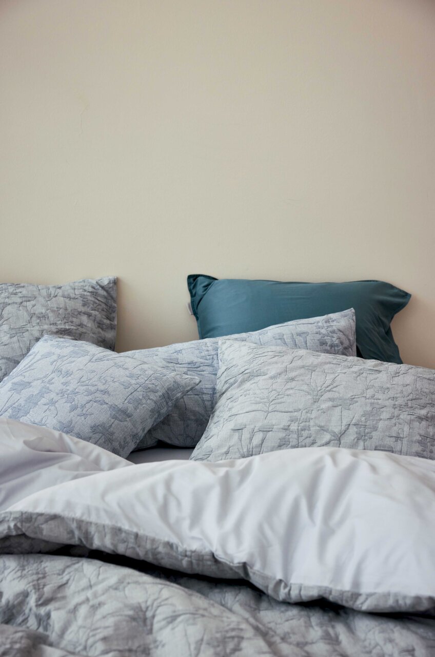 Bettwäsche-Set mit floralem Muster in Grautönen, bestehend aus mehreren Kissen und einer Decke, aus der Perspektive von der Seite fotografiert.