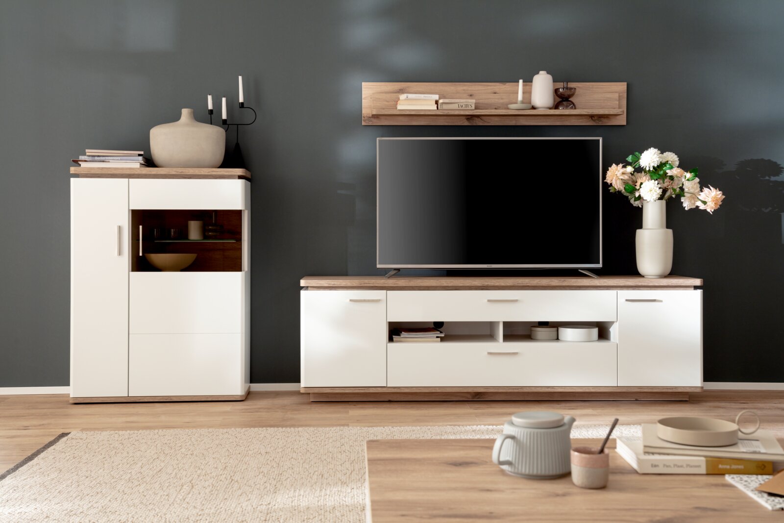 Modernes TV-Wohnwand-Set in Weiß mit Holzoptik (Lowboard, Highboard und Wandregal) in einem Wohnzimmer, frontal aus Augenhöhe fotografiert.