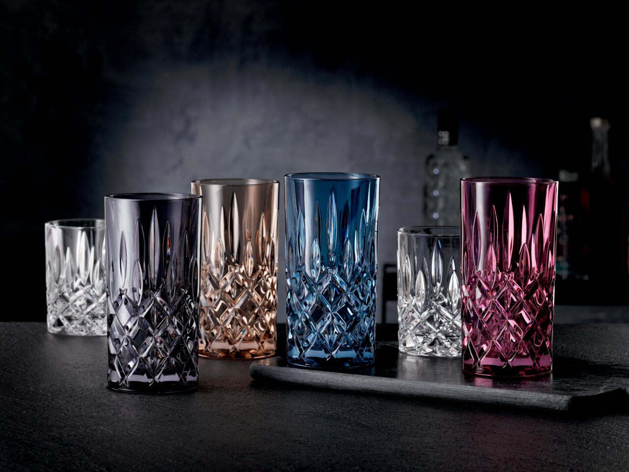 Verschiedene farbige Longdrinkgläser aus Kristallglas in einer eleganten Anordnung auf einem dunklen Tisch, seitliche Perspektive.