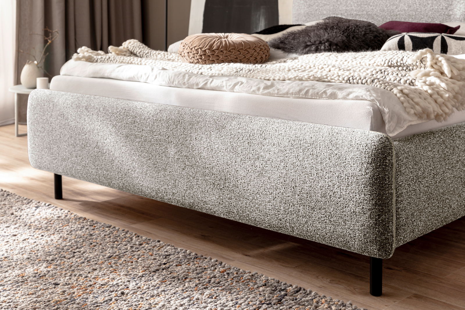 Seitliche Perspektive eines modernen Polsterbetts mit grauem Stoffbezug, dekoriert mit Kissen und einer Decke.