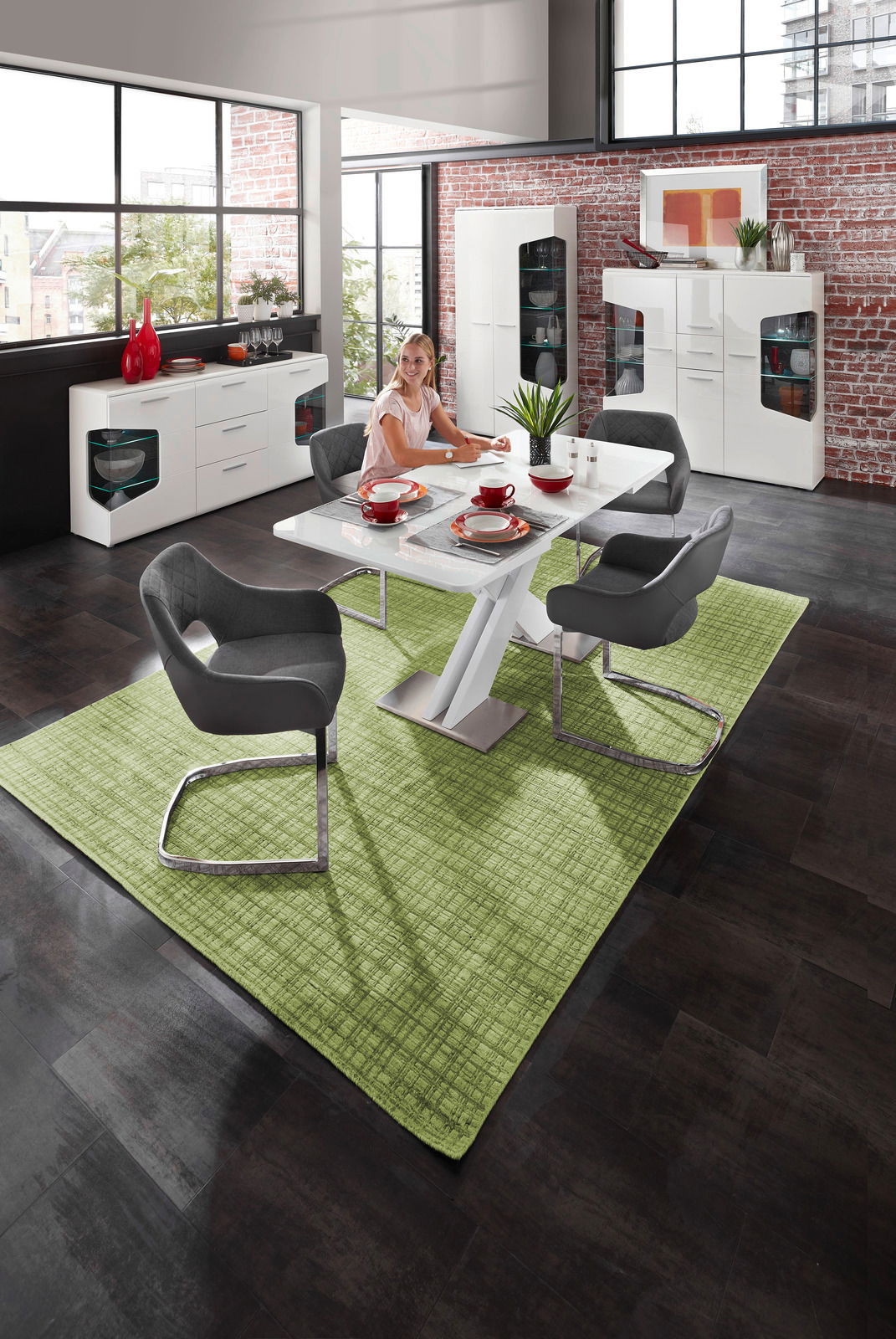 Moderne Esszimmer-Szene mit einem weißen Funktions-Esstisch, umgeben von grauen Armlehnstühlen auf einem grünen Teppich. Im Hintergrund sind ein Sideboard und eine Highboard-Vitrine vor einer Backsteinwand zu sehen. Perspektive: Blick von oben auf den Tis