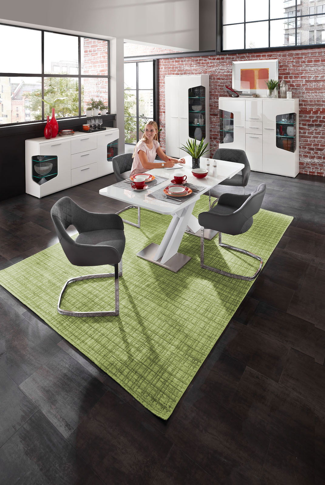 Moderne Esszimmer-Szene mit einem weißen Funktions-Esstisch, umgeben von grauen Armlehnstühlen auf einem grünen Teppich. Im Hintergrund sind ein Sideboard und eine Highboard-Vitrine vor einer Backsteinwand zu sehen. Perspektive: Blick von oben auf den Tis