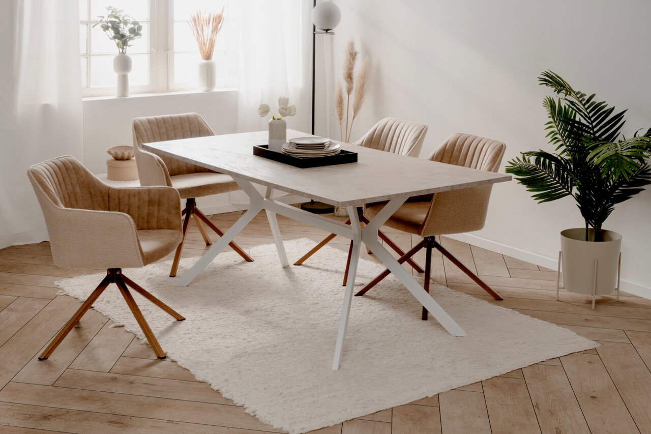 Moderner Esstisch mit heller Tischplatte und weißen Metallbeinen, umgeben von vier beigen Stühlen auf einem hellen Teppich in einem hellen Raum mit Pflanzen und Dekorationen. Perspektive von schräg oben.