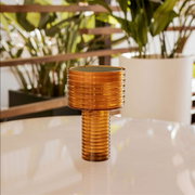 Een geribbelde tafellamp van amberkleurig glas met een cilindrische kap en voet, vooraanzicht vanuit een licht verhoogd perspectief op een witte tafel, met op de achtergrond onscherpe planten.