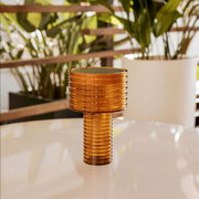 Gerippte Tischlampe aus bernsteinfarbenem Glas mit zylindrischem Schirm und Fuß, frontal aus leicht erhöhter Perspektive auf einem weißen Tisch, im Hintergrund unscharfe Pflanzen.