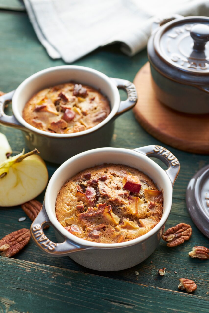 Staub Cocotte Set 4-tlg. Zwei graue Mini-Cocottes mit gebackenem Dessert, umgeben von Apfelstücken und Nüssen, aus der Vogelperspektive fotografiert.