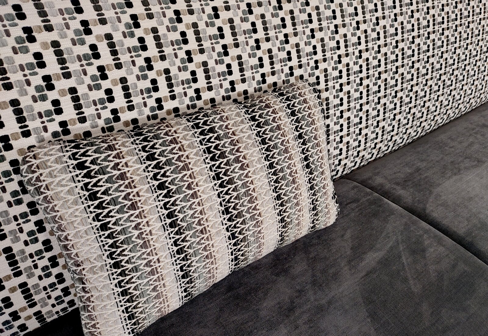Nahaufnahme in schräger Seitenperspektive: dekoratives Sofa-Kissen mit gewebtem Zickzack- und Streifenmuster in Beige, Grau und Schwarz auf grauer Sitzfläche vor gemusterter Rückenlehne