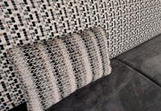 Nahaufnahme in schräger Seitenperspektive: dekoratives Sofa-Kissen mit gewebtem Zickzack- und Streifenmuster in Beige, Grau und Schwarz auf grauer Sitzfläche vor gemusterter Rückenlehne