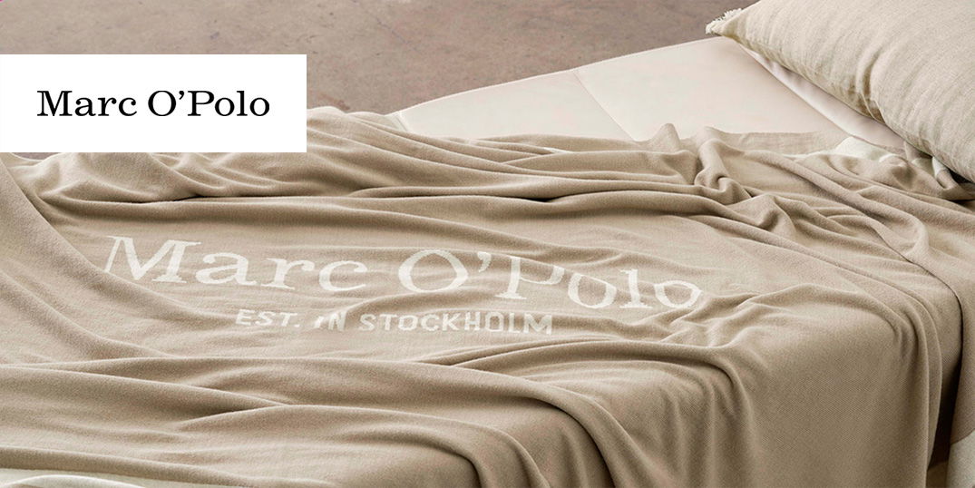 Beiges Plaid mit Marc O’Polo-Logo auf einem Bett mit Kissen.
