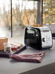 Schwarzer KitchenAid 2-Scheiben-Toaster in moderner Küche, seitliche Perspektive, mit Toastscheiben und Frühstückszubehör.