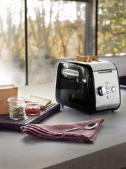 Schwarzer KitchenAid 2-Scheiben-Toaster in moderner Küche, seitliche Perspektive, mit Toastscheiben und Frühstückszubehör.