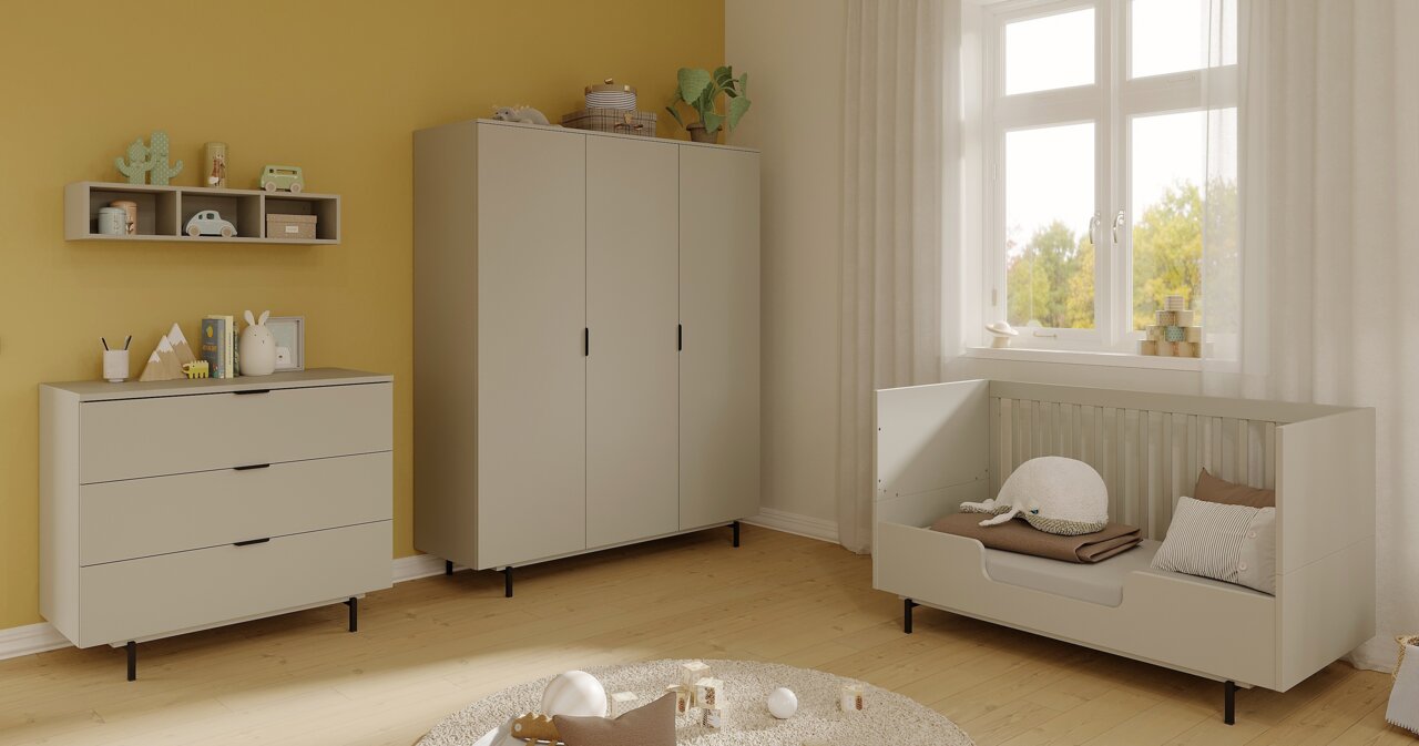 Kinderzimmer mit einer modernen, grauen Kommode, die von vorne zu sehen ist. Die Kommode hat drei Schubladen und steht auf schwarzen Beinen. Sie ist dekoriert mit Spielzeug und Büchern. Im Hintergrund sind ein Kleiderschrank und ein Kinderbett zu sehen.