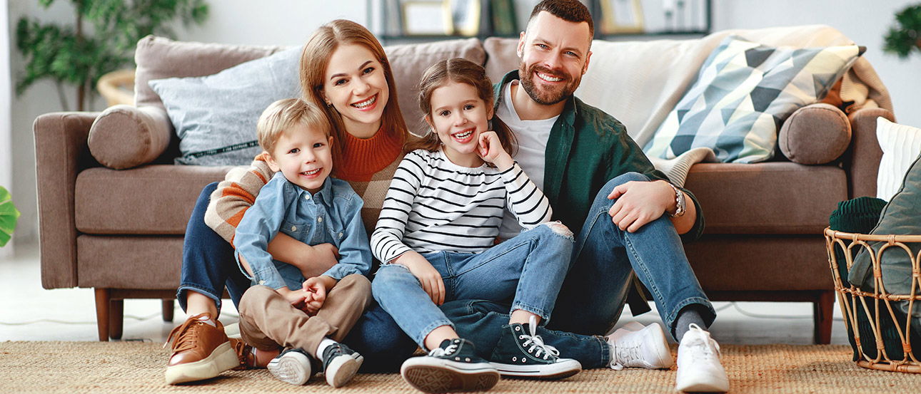 Familie sitzt zusammen auf dem Boden im Wohnzimmer vor einem braunen Sofa.