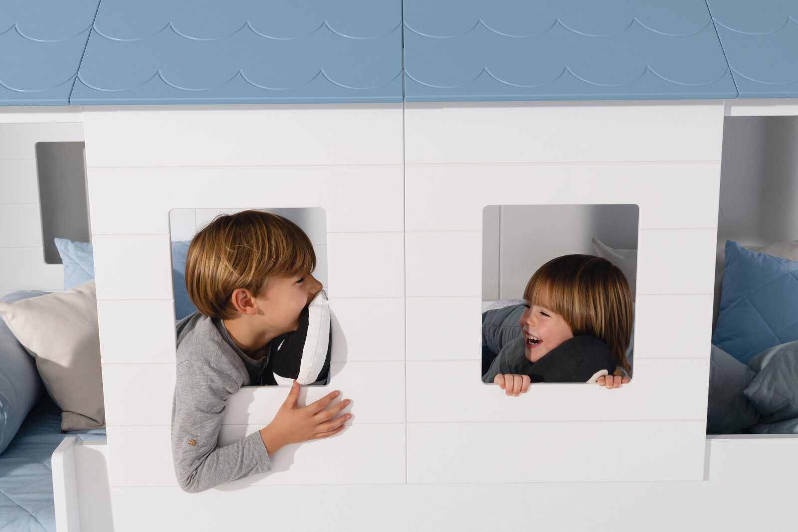 Zwei Kinder spielen fröhlich in einem weißen Fensterpaneele-Set mit blauen Dachpaneelen. Die Perspektive zeigt die Vorderansicht des Produkts.