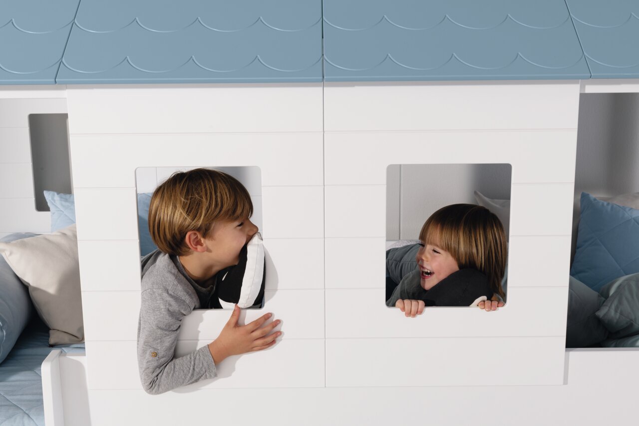Zwei Kinder spielen fröhlich in einem weißen Fensterpaneele-Set mit blauen Dachpaneelen. Die Perspektive zeigt die Vorderansicht des Produkts.