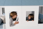 Zwei Kinder spielen fröhlich in einem weißen Fensterpaneele-Set mit blauen Dachpaneelen. Die Perspektive zeigt die Vorderansicht des Produkts.