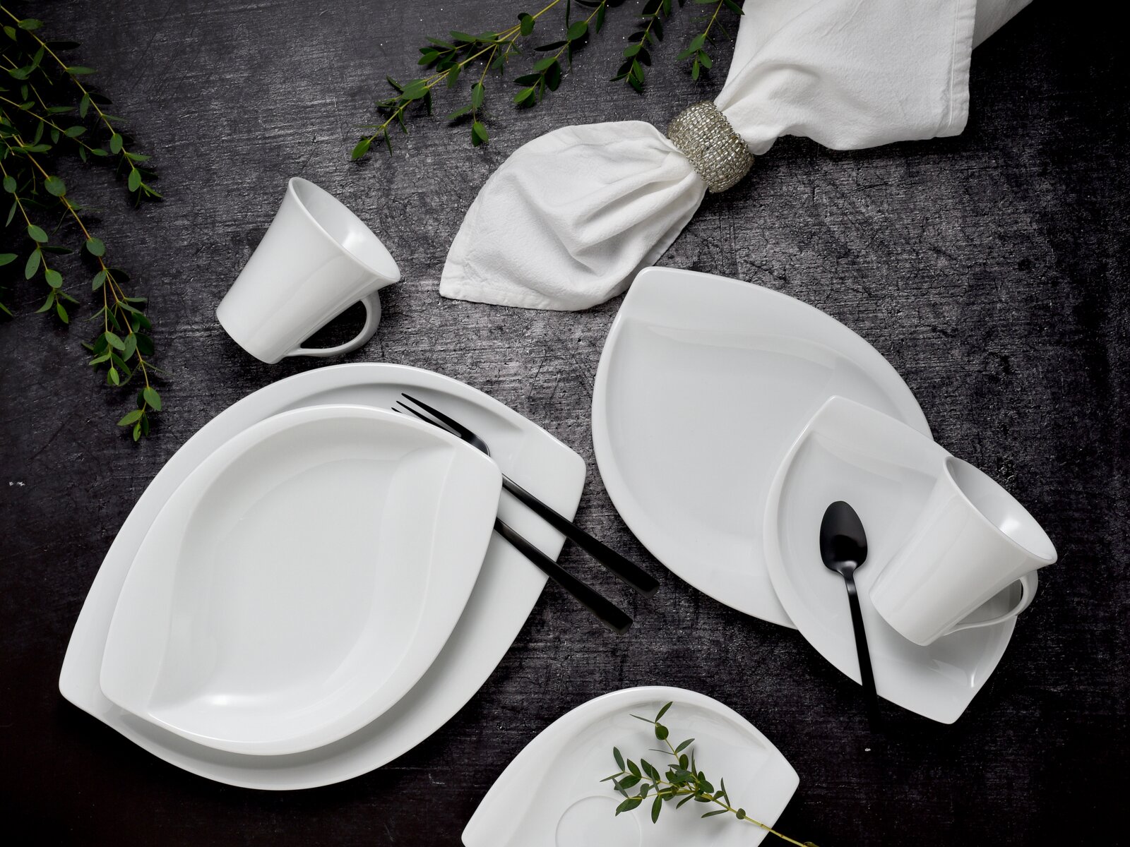 Weißes Porzellan-Geschirrset mit Tellern, Schalen und Tassen auf dunklem Untergrund, dekoriert mit grünem Zweig und weißer Serviette mit silbernem Ring, fotografiert in Draufsicht (Top-Down)