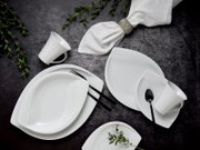Weißes Porzellan-Geschirrset mit Tellern, Schalen und Tassen auf dunklem Untergrund, dekoriert mit grünem Zweig und weißer Serviette mit silbernem Ring, fotografiert in Draufsicht (Top-Down)