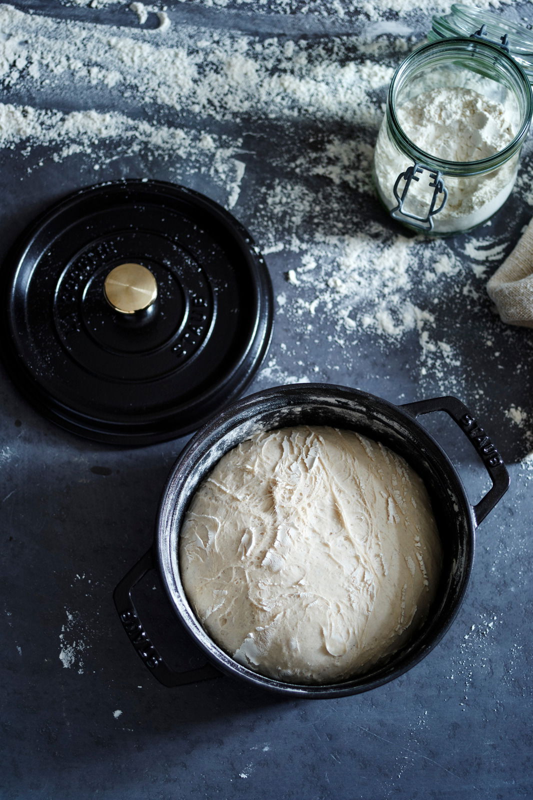 Runde schwarze Cocotte mit Teig, Deckel daneben, von oben fotografiert, auf bemehlter Oberfläche.